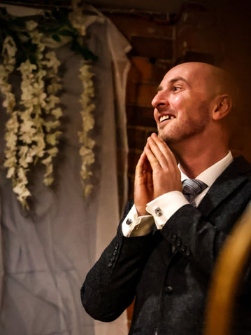 Smiling man in suit clasps hands, looking up, with floral background setting.