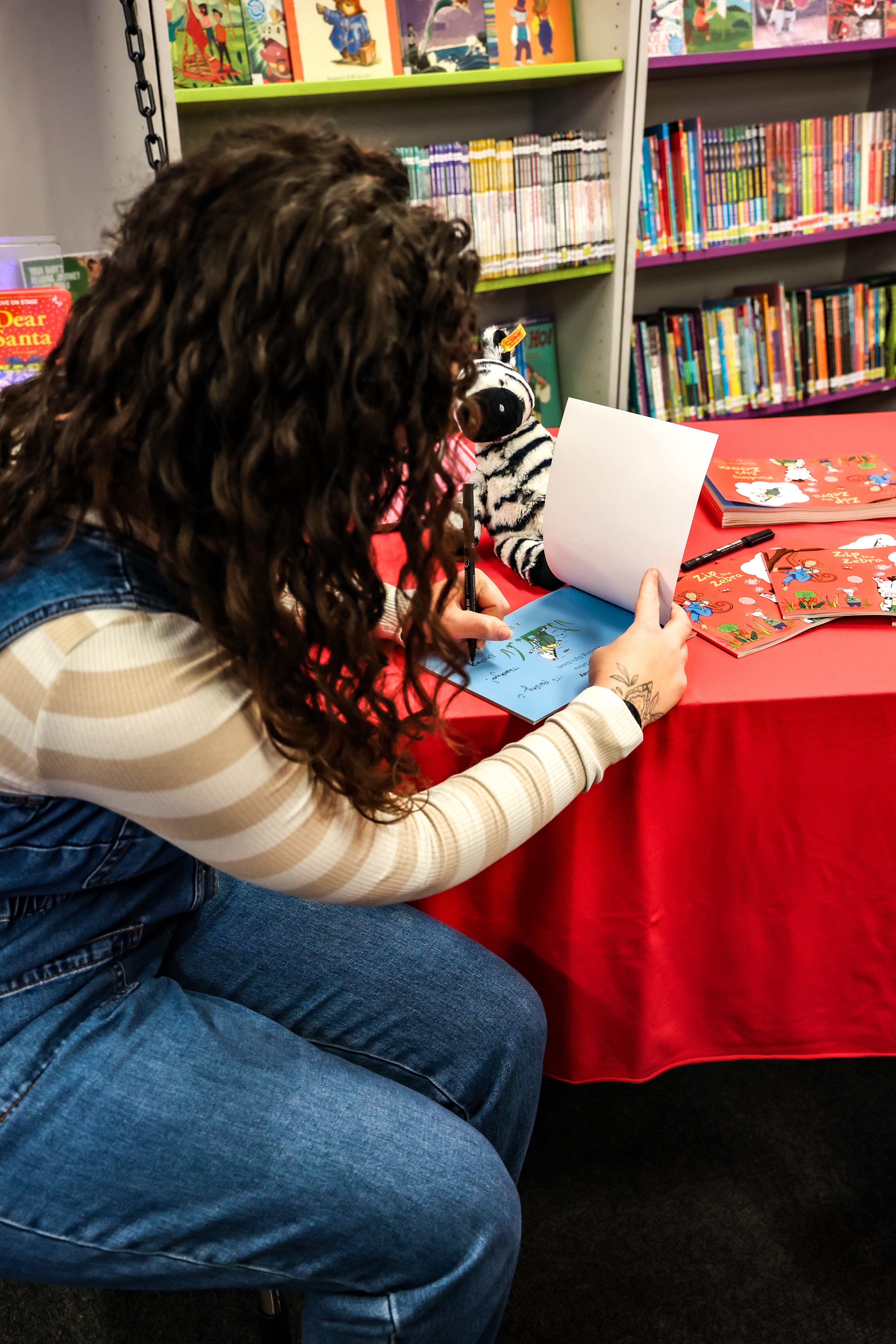 Woman signs a book whilst leaning on a red table in library.