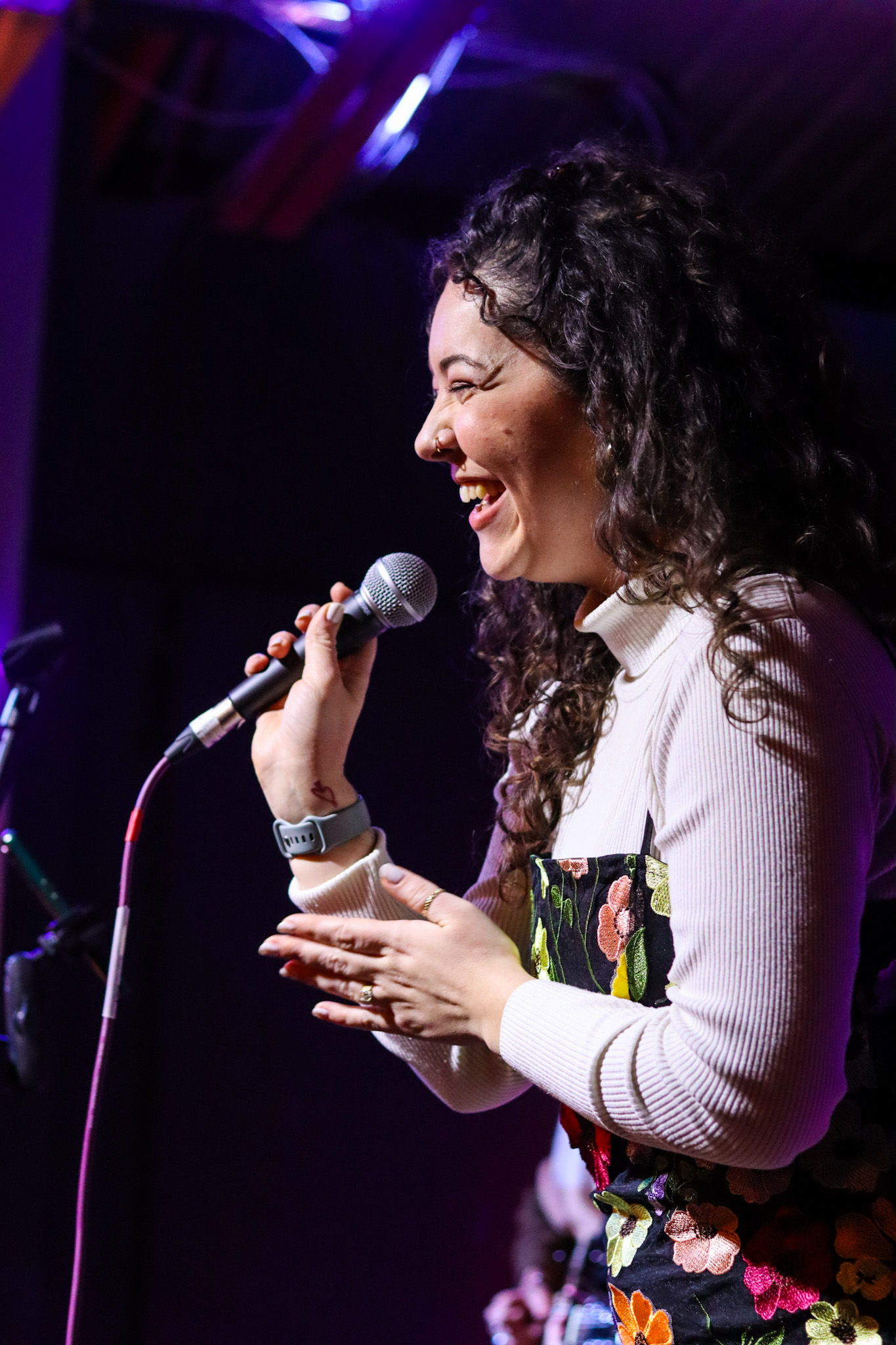 Smiling woman sings into microphone on stage with colorful floral dress.