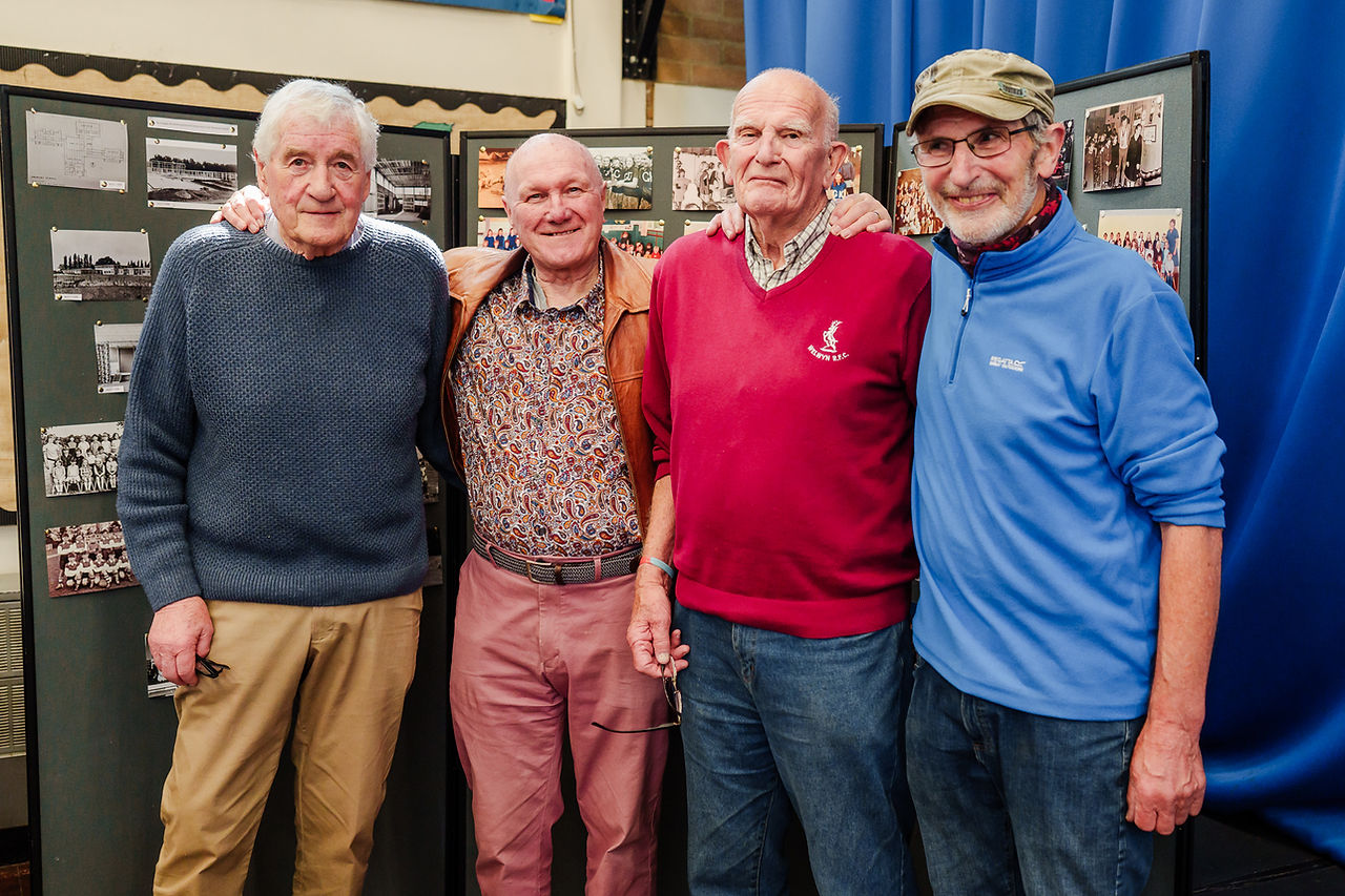 Four older gentlemen pose for a photo in front of a board displaying old photographs.