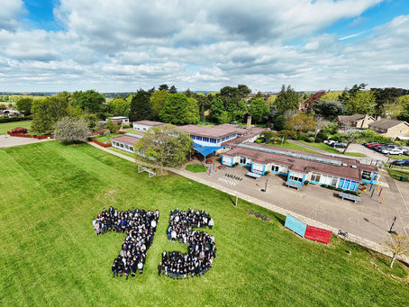 Aerial view of people forming "75" on a green field beside a school building under a cloudy sky, celebrating an event with a joyful mood.
