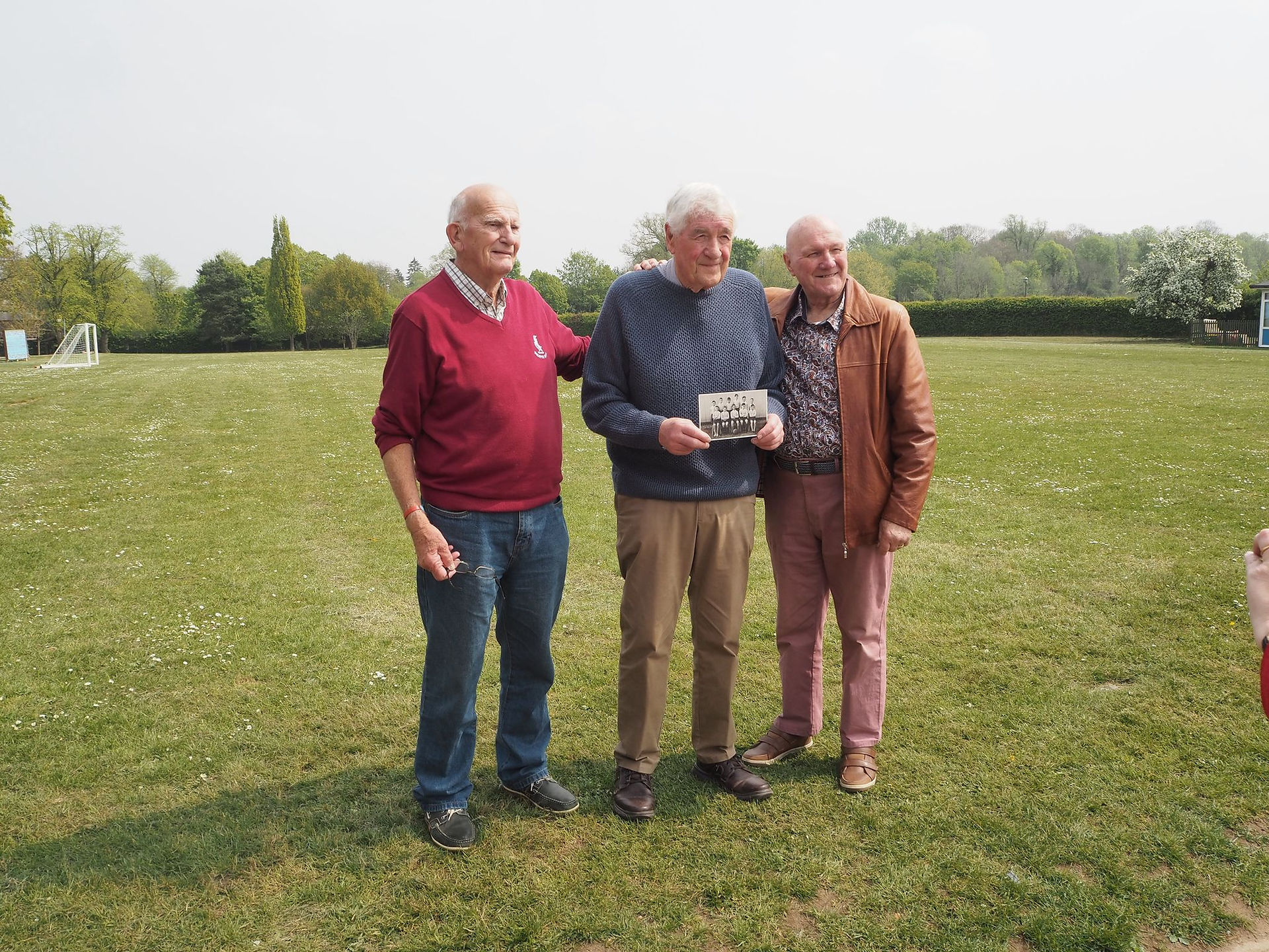 three elderly gentlemen stand together on a school field holding a black and white school photo