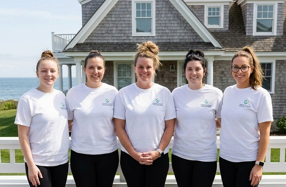 Five women in white shirts stand smiling outdoors by a beach house. Ocean visible in the background. Shirts say "Green Clean Innovations."