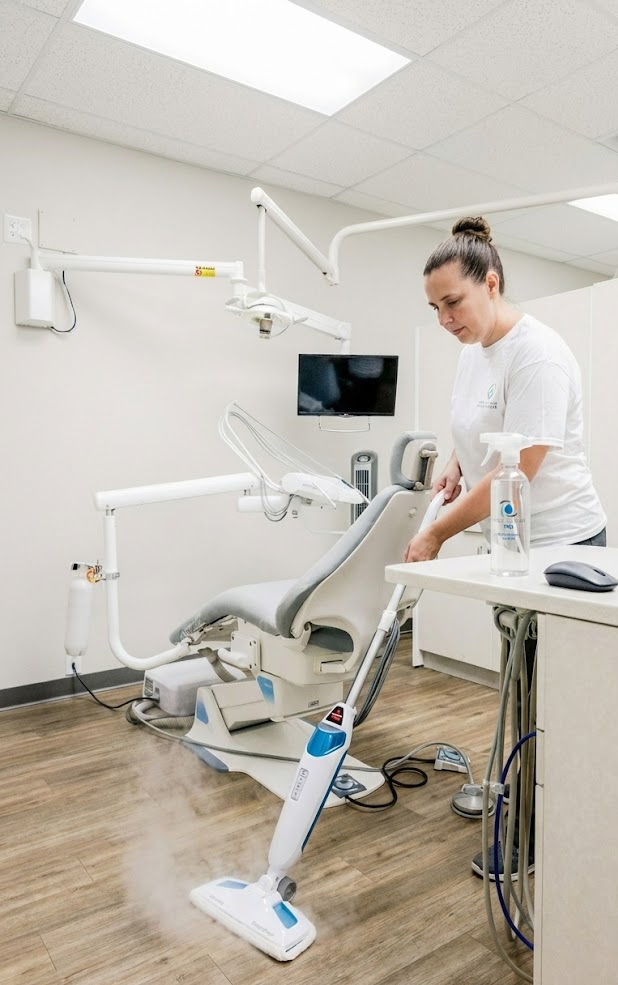 A woman cleans a dental office with a steam mop. The setting is a bright, modern clinic with a dental chair and equipment. Clean and tidy mood.