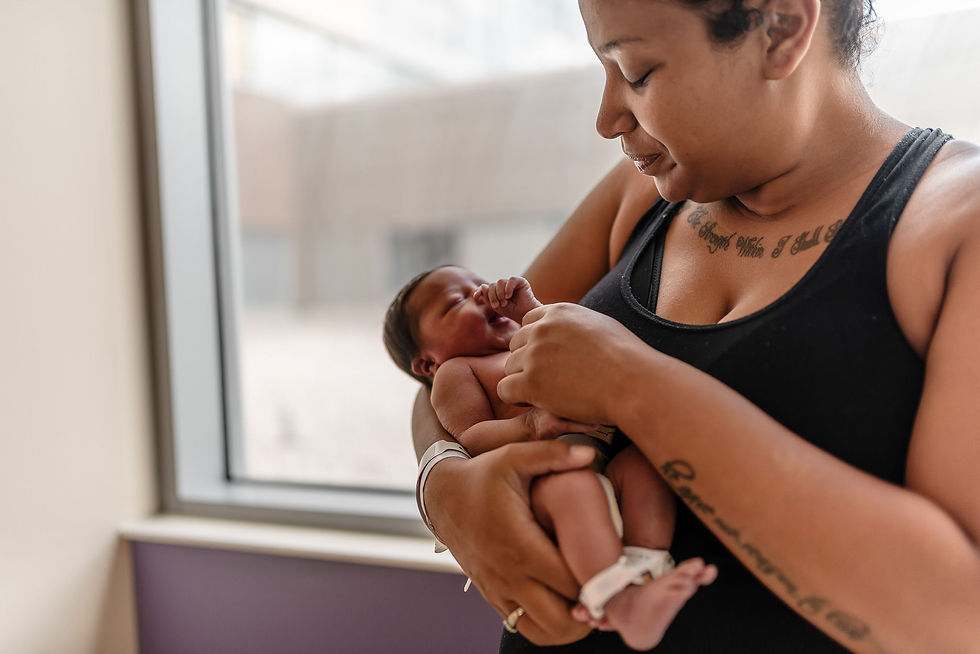 New mother holding newborn baby near hospital window during postpartum recovery after birt