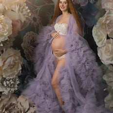 Fine art pregnancy photo of a mother-to-be in flowing lavender robes, sitting in a flower-draped archway. Studio photo with dreamy tones and a soft magical mood.