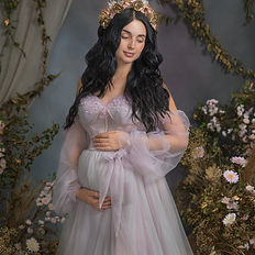 Elegant fine art maternity portrait of a dark-haired pregnant woman in a soft lavender gown, holding her belly with both hands. She stands in front of a botanical-inspired studio background with trailing green vines and delicate pink florals, evoking serenity, grace, and anticipation. Captured in Michaela Ďurišová’s signature fine art style in Bratislava.
