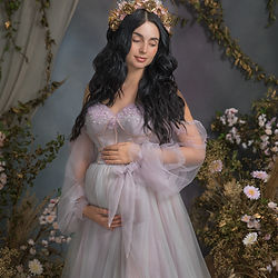 Elegant fine art maternity portrait of a dark-haired pregnant woman in a soft lavender gown, holding her belly with both hands. She stands in front of a botanical-inspired studio background with trailing green vines and delicate pink florals, evoking serenity, grace, and anticipation. Captured in Michaela Ďurišová’s signature fine art style in Bratislava.