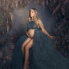 Maternity fine art photo of a woman wearing a flowing teal gown, seated gracefully among pastel flowers. Stylized scene with soft light and floral storytelling.