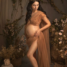 Fine art photo of a pregnant woman styled in bronze-golden tones, standing gracefully in warm cinematic lighting. Surrounded by earthy textures and dried florals, this portrait evokes a timeless, goddess-like presence. A celebration of maternal strength and elegance, photographed in a richly styled studio setting.
