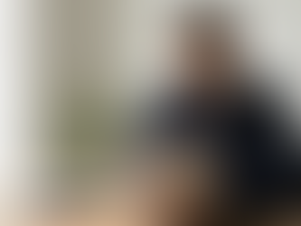 A man reads a book titled "L'Étranger" at a wooden table. A notebook lies nearby. A green plant is in the background, setting a calm mood.