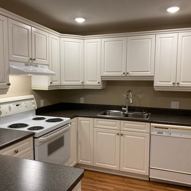 Empty, freshly cleaned white kitchen with peninsula and dark gray laminate countertops in a rental suite.