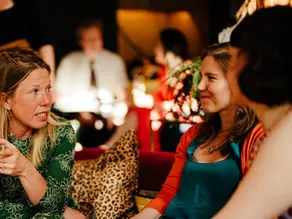People sitting together indoors having a warm conversation during a community social gathering, with a woman speaking while others listen and smile.