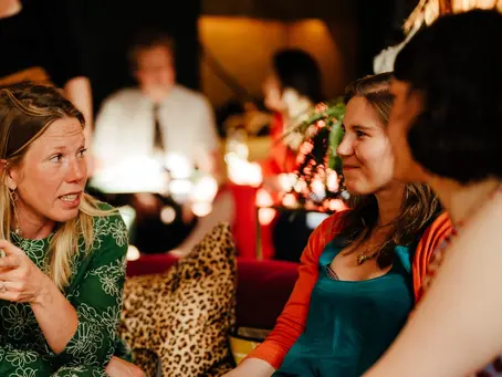 People sitting together indoors having a warm conversation during a community social gathering, with a woman speaking while others listen and smile.