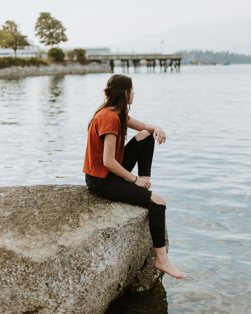 A young woman sitting on a rock at the water's edge looking into the distance