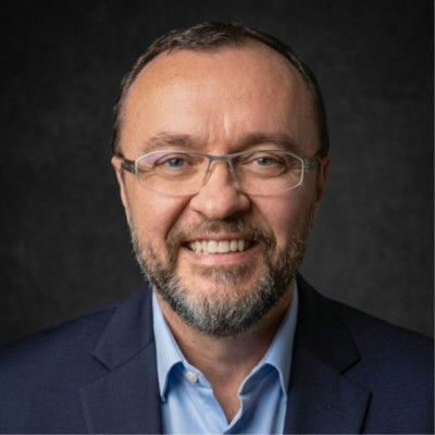 Smiling middle-aged man with glasses and a beard in a navy suit, posed against a dark studio background.