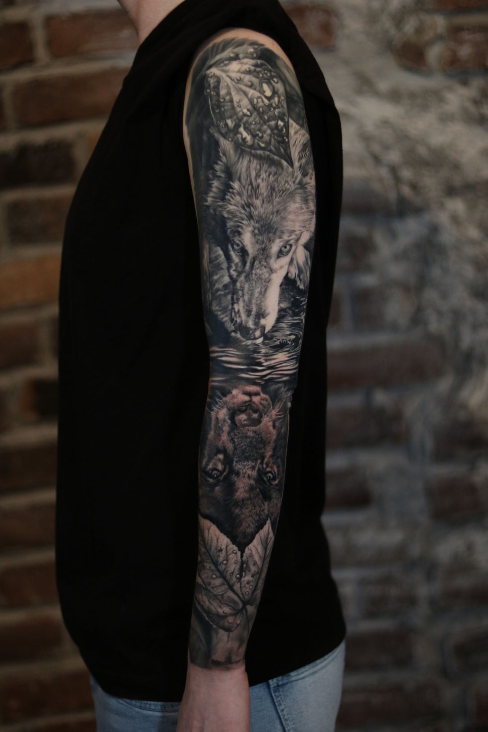 Black and grey realism sleeve tattoo of a wolf reflected in water as a darker version of itself, surrounded by detailed leaves.