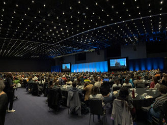Wide angle ballroom view of attendees eating lunch at round tables facing general session stage
