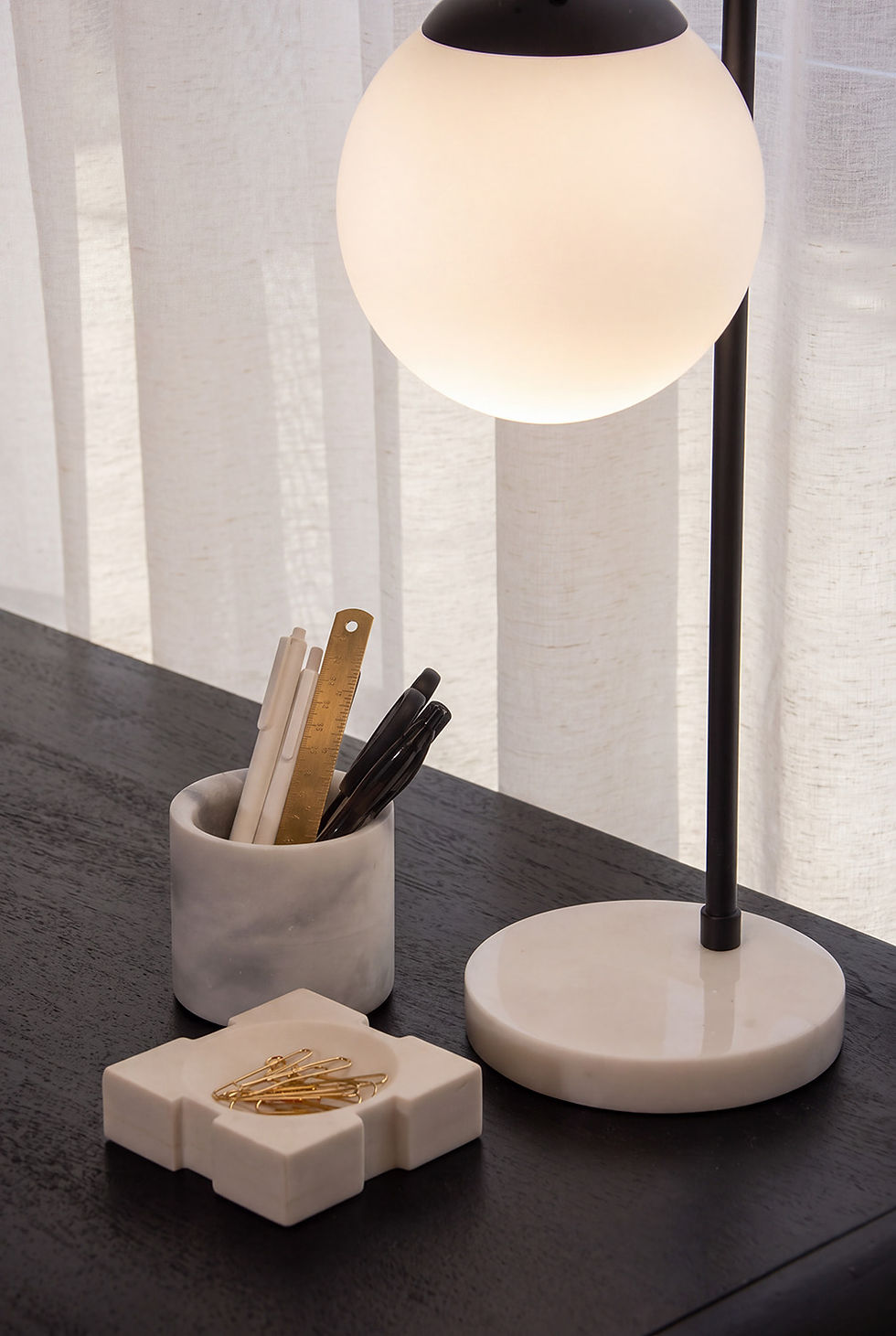 Close-up of a frosted globe desk lamp with a white marble base beside a marble pen cup holding white pens, a brass ruler and black scissors, and a marble tray of gold paperclips