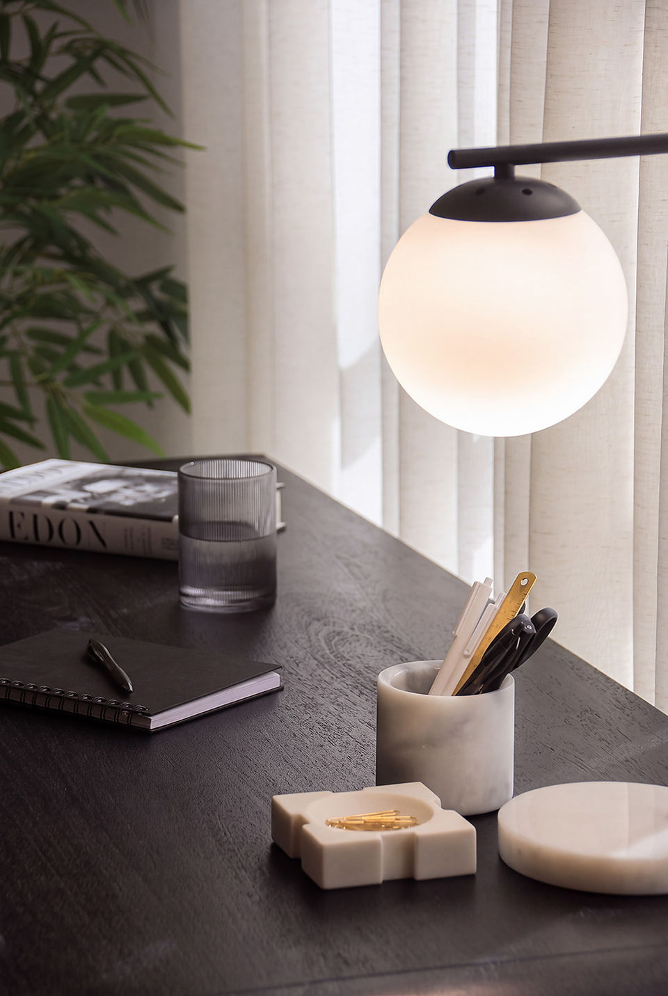 Desk detail with a frosted globe lamp, marble pen cup and paperclip tray, ribbed glass, black notebook and Avedon book, with bamboo leaves in the foreground