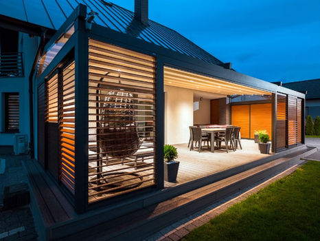 Dark aluminium pergola wrapping round a house corner at dusk with timber-effect louvred shutters and dining table