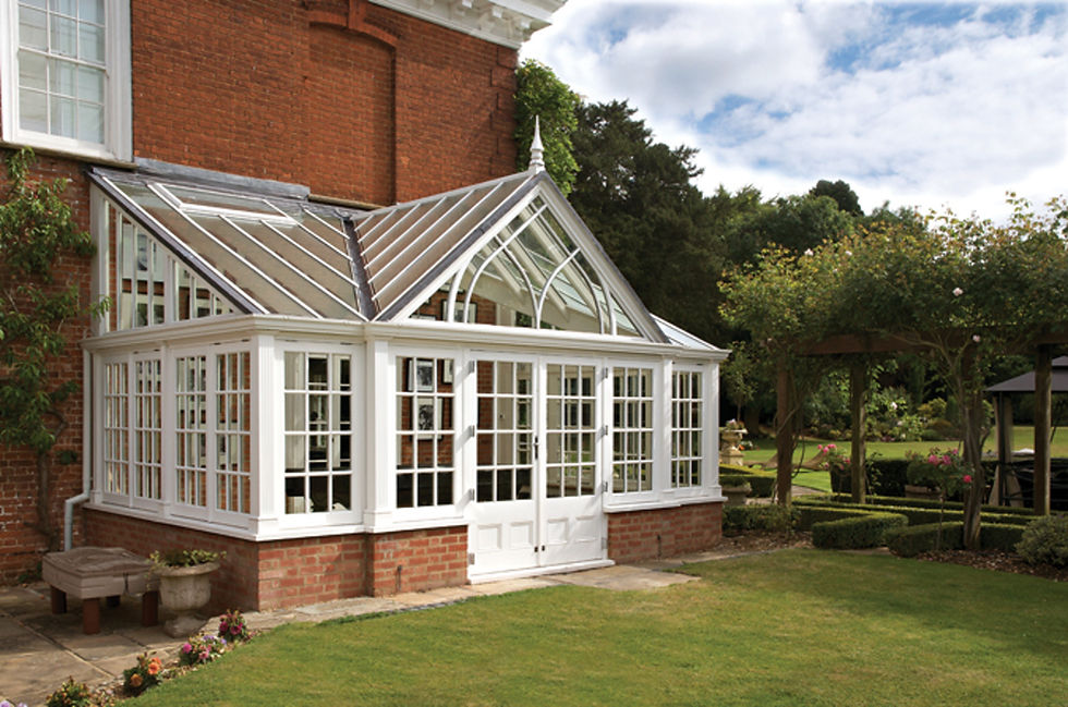 White painted timber gable-fronted conservatory with a pitched glass roof on a brick base beside a red-brick house