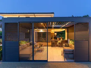 Anthracite louvred-roof pergola with sliding louvred side shutters, lit at dusk over a garden kitchen