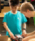 Two boys work on a robot in a classroom. One wears a blue shirt with text; the other wears green. They're focused and engaged with the task.