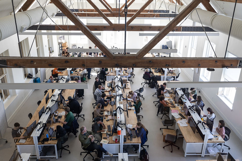 Photo taken from above of a large room with wooden rafters and hanging light fixtures over 6 double sided bench desks with computers.