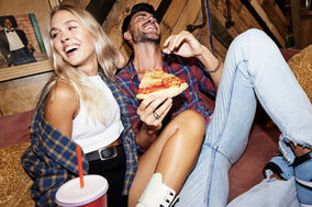 Food lifestyle photography of couple eating pizza and drinking soda on couch, playful casual home setting