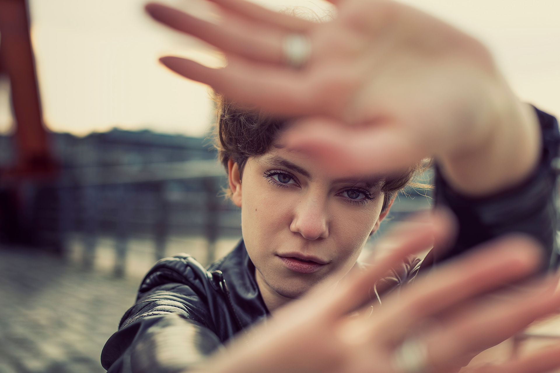 woman in a black leather jacket covering camera lens with her hands Urban background is slightly blurred, giving a moody vibe.