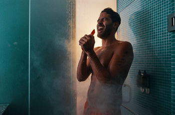 A man stands in a steamy shower, water cascading down, creating a misty atmosphere around him. 