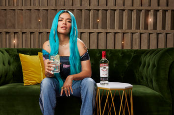 Karol G seatting on a sofa holding a glass of vodka