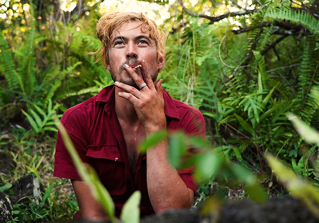 A dirt streaked man in his 30's sits among the ferns. He smokes on a cigarette and smiles a smirk at the camera.