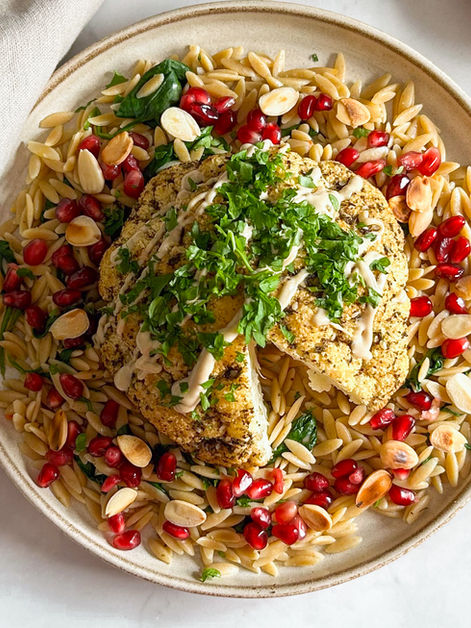 a plate of orzo with a piece of roasted cauliflower and tahini sauce, nuts and pomegranate with greens on top