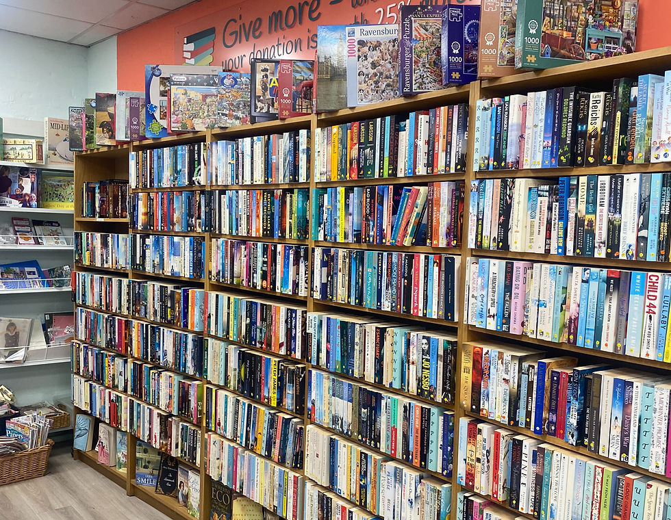 Large wall of second-hand books displayed in a charity shop, with shelves filled with fiction and non-fiction titles and jigsaw puzzles on top.