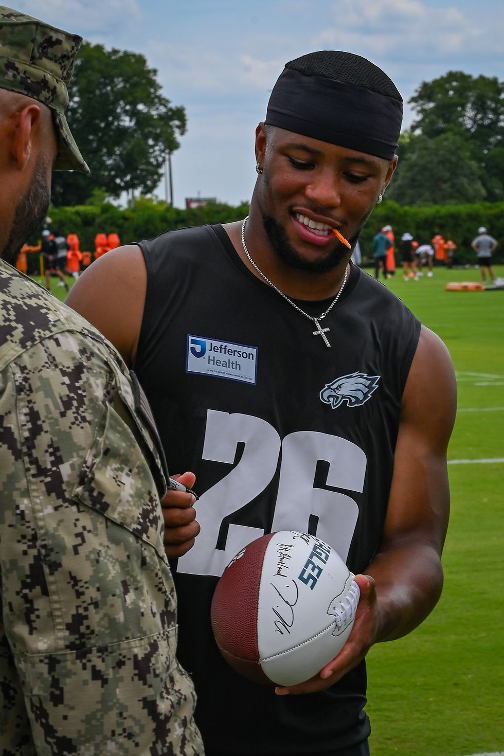 Saquon Barkley in a black Philadelphia Eagles No. 26 training-camp jersey signing a football for a service member at the NovaCare Complex on July 26, 2025.