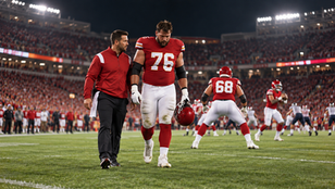Editorial illustration of a fictional red-uniformed offensive tackle leaving the field beside a trainer while his replacement prepares