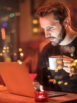 Man in cafe, focused on laptop, holds white mug. Warm lighting with bokeh effect in background. Cozy, contemplative mood.