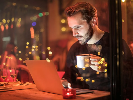 Man in cafe, focused on laptop, holds white mug. Warm lighting with bokeh effect in background. Cozy, contemplative mood.