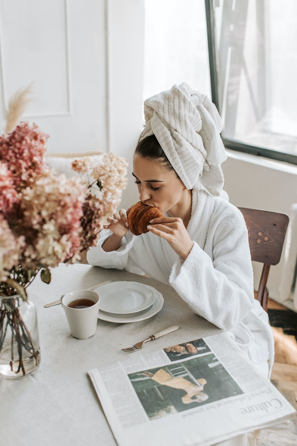 Mulher vestida com roupão de banho e comendo croissant e tamando café em casa