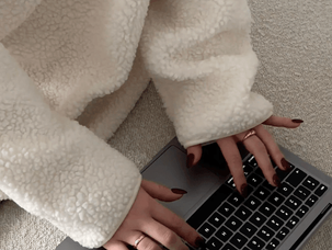 Pessoa em casaco bege digita em laptop cinza sobre sofá claro. Mãos bem cuidadas, unhas escuras. Ambiente tranquilo e minimalista.