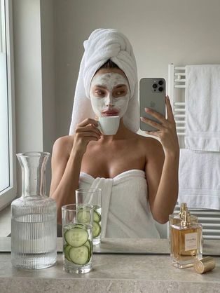 Mulher com máscara facial e toalha na cabeça, tira selfie enquanto bebe café. Em volta, jarra de água, vidros com pepino e perfume Chanel.