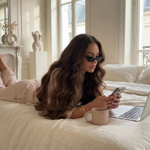 Mulher de óculos escuros deitada na cama bege, mexe no celular ao lado de um laptop e uma caneca, em quarto elegante e claro.