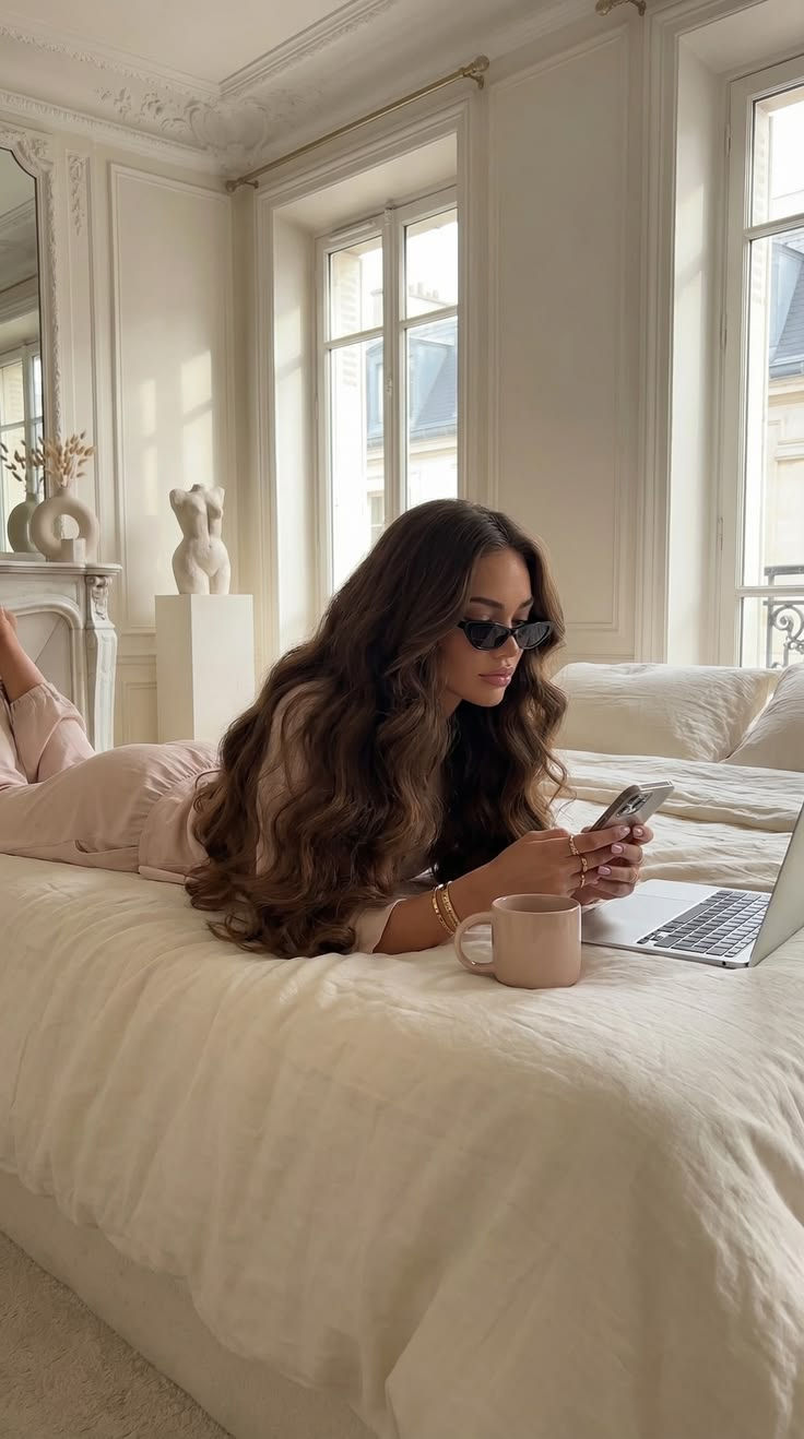 Mulher de óculos escuros deitada na cama bege, mexe no celular ao lado de um laptop e uma caneca, em quarto elegante e claro.