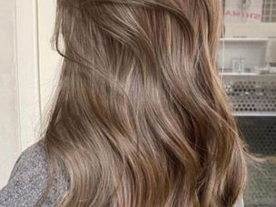 Cabelo castanho claro ondulado visto de trás, em salão, com suéter cinza e pôster ao fundo.