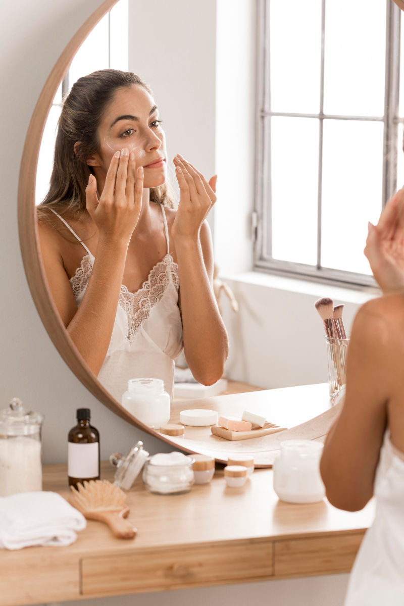 Mulher aplica creme no rosto diante do espelho, em penteadeira clara com produtos de skincare e luz suave.