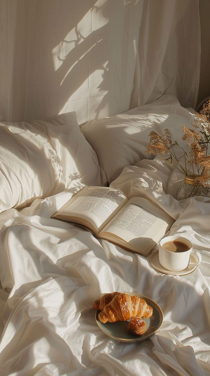 Cama com lençóis brancos, livro aberto, xícara de café e croissant. Luz suave entra pela janela, criando sombras calmantes.