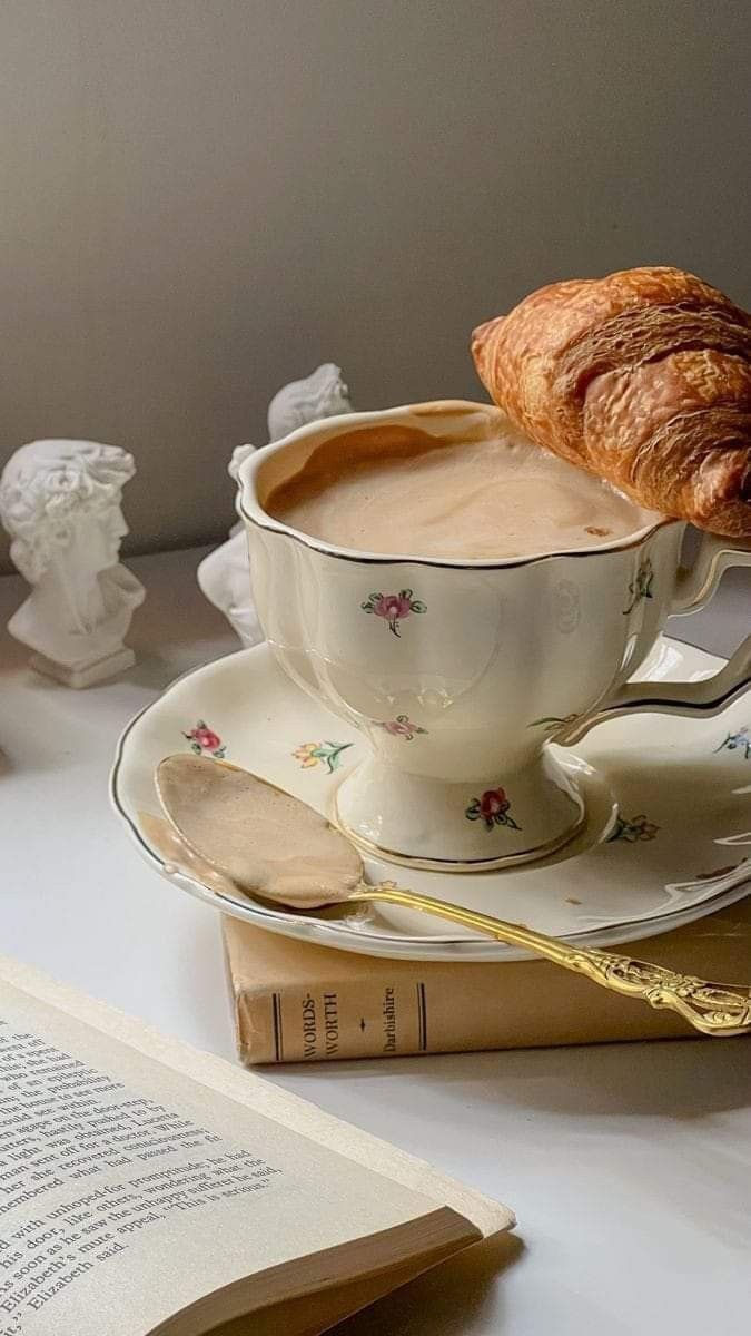 Xícara floral de chá com croissant, colher dourada e livros; na lombada lê-se Words Worth, clima calmo e aconchegante.
