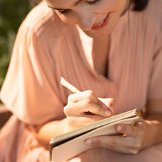 Mulher sorridente escreve em um caderno ao ar livre, em luz dourada, com blusa rosa.